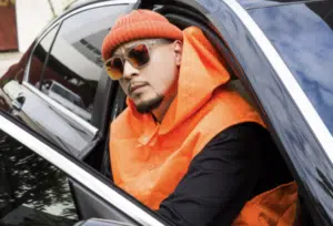 A man wearing an orange beanie, orange vest, and sunglasses leans out of the window of a black car.